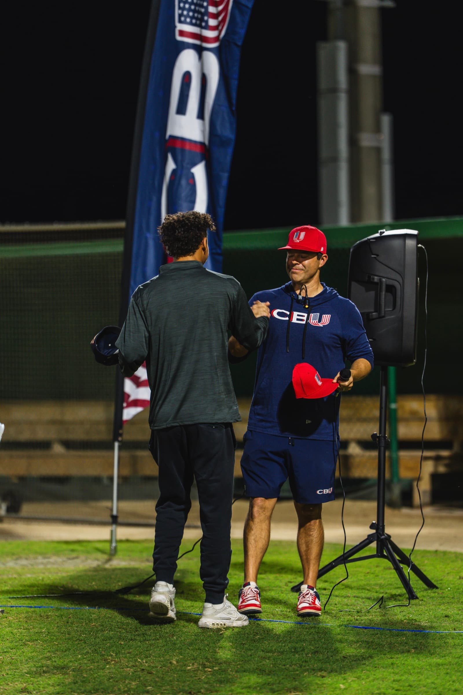 Coach handing red hat to player on the field at night