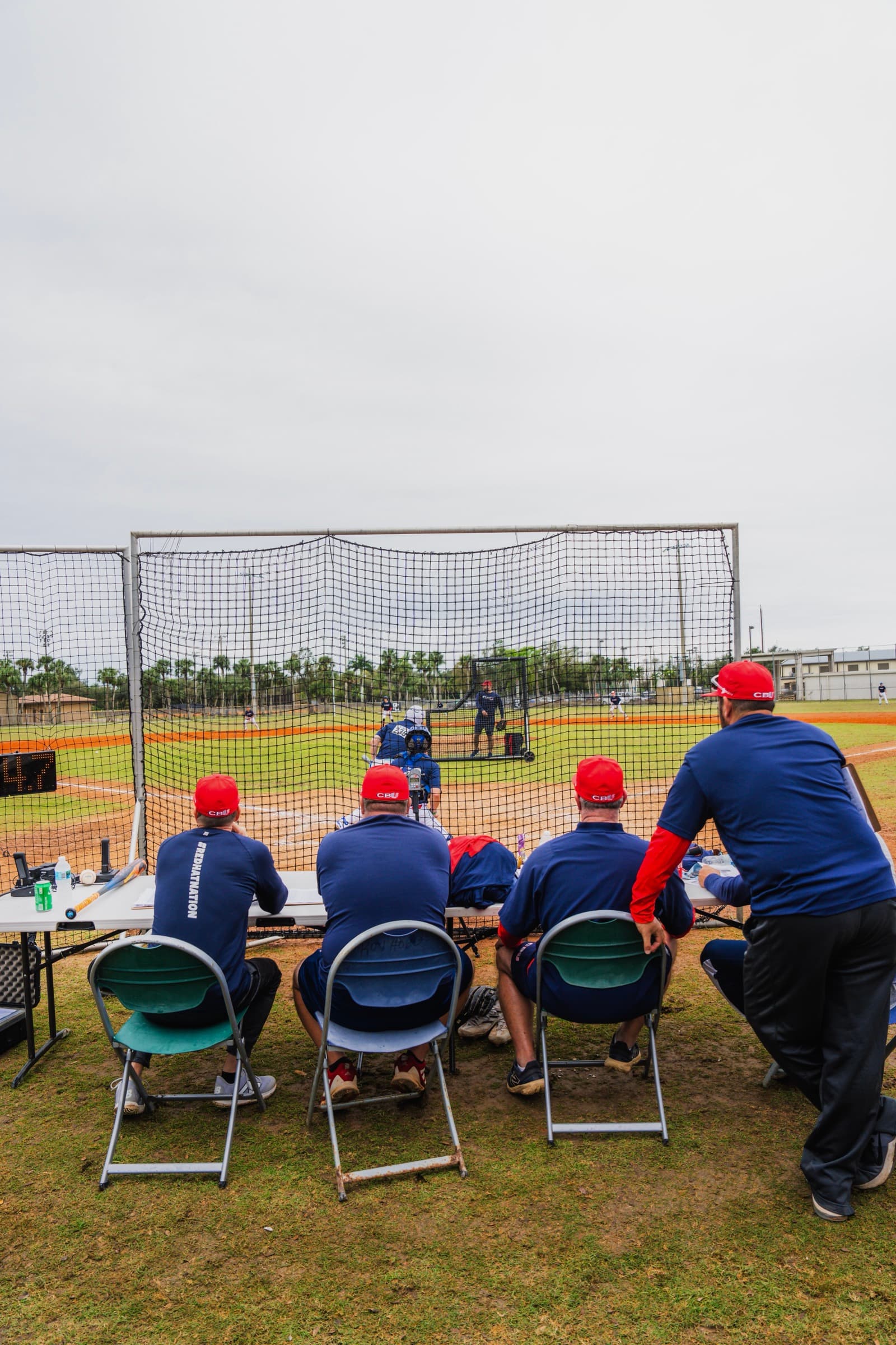 Row of coaches in red hats evaluating players behind batting cage